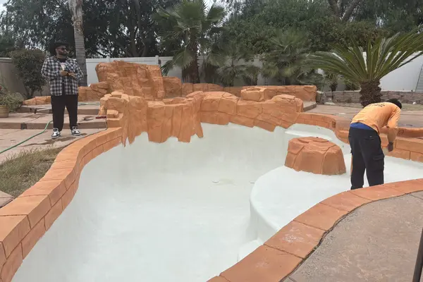 Ground-level view of the pool with fresh white plaster, sculpted rockwork around the edge and a worker finishing the surface