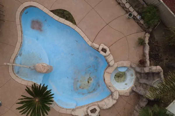 Aerial view of a worn freeform pool with faded blue plaster and dark staining across the floor